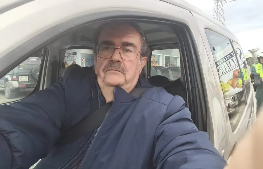 Hombre con gafas y bigote, sentado al volante de una furgoneta y haciéndose una foto con el brazo extendido; al fondo se ve la carretera y un agente de Tráfico junto a una moto.