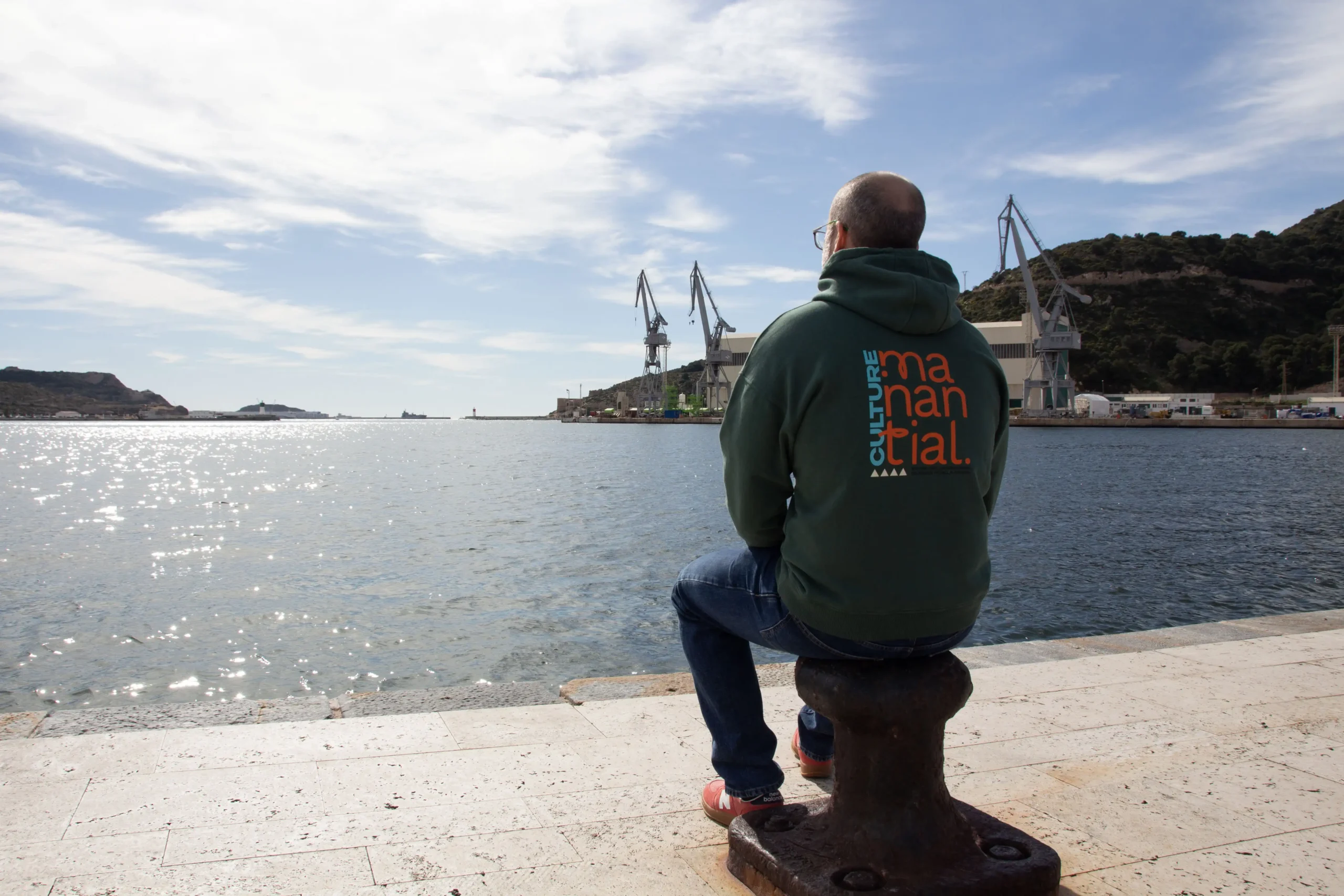 Hombre sentado frente al puerto de Cartagena contemplando el mar, con grúas industriales al fondo, vistiendo sudadera verde de Manantial Wear con diseño tipográfico en la espalda.