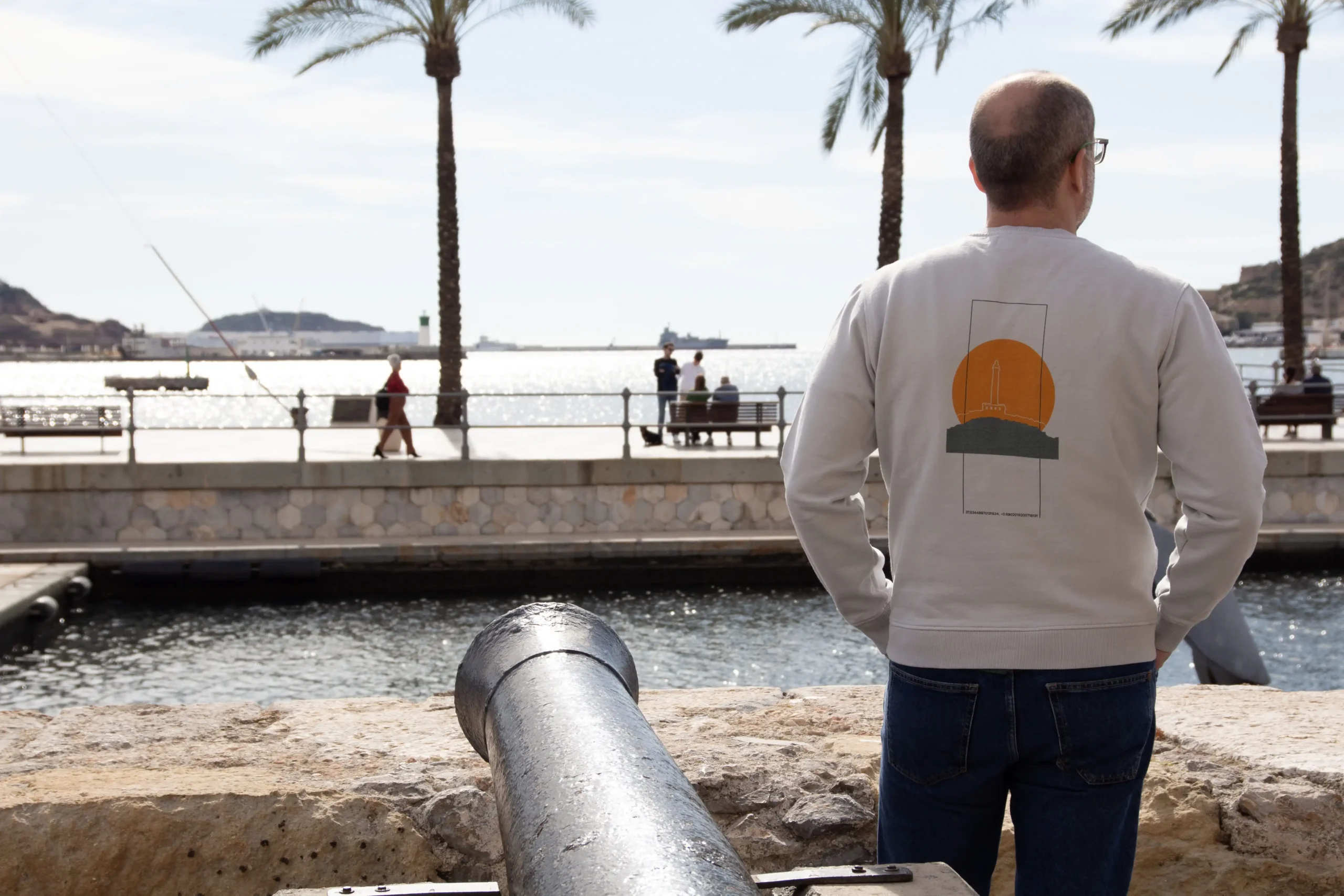 Hombre de espaldas contemplando el puerto de Cartagena junto a un cañón histórico, con palmeras y el mar al fondo, vistiendo una sudadera de Manantial Wear con diseño de faro y sol mediterráneo.