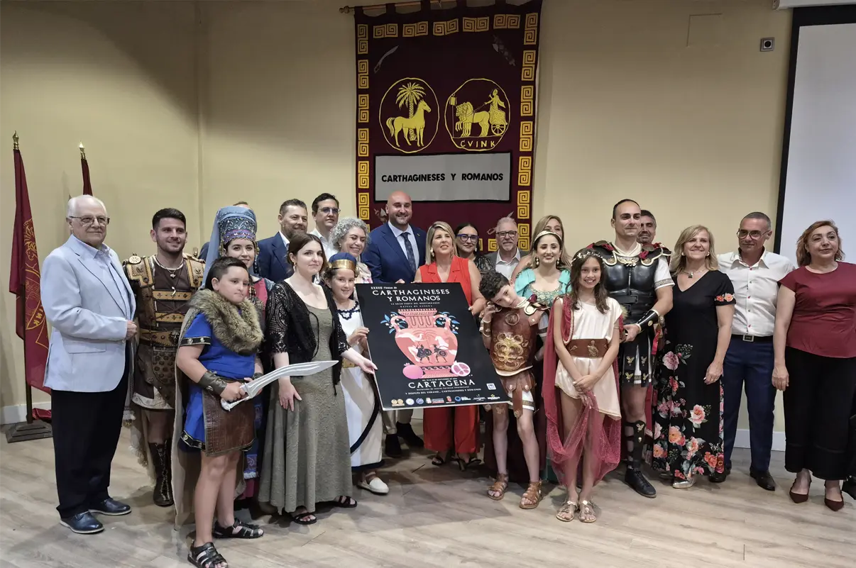Autoridades, representantes de la Federación de Tropas y Legiones, personajes festeros infantiles y adultos, y la autora Natalia Lucas posan junto al cartel oficial de la XXXVII edición de Carthagineses y Romanos durante su presentación en Cartagena.